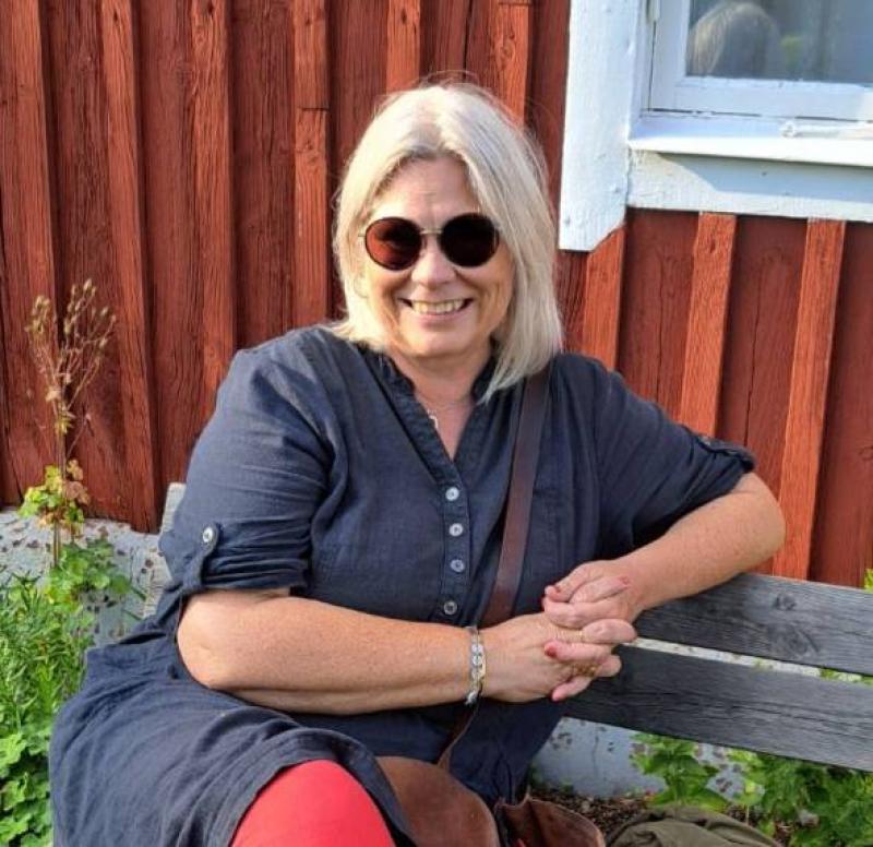 En kvinna med blont hår och solglasögon sitter på en bänk framför en röd träbyggnad och ler. Hon är klädd i en mörk klänning och röda byxor.