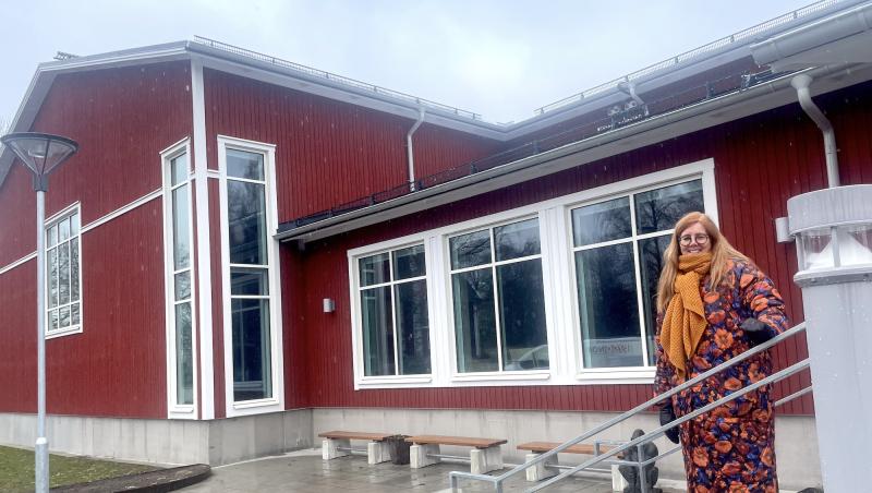 En kvinna i en blommig klänning och orange halsduk ler utanför en röd byggnad med en skylt märkt 