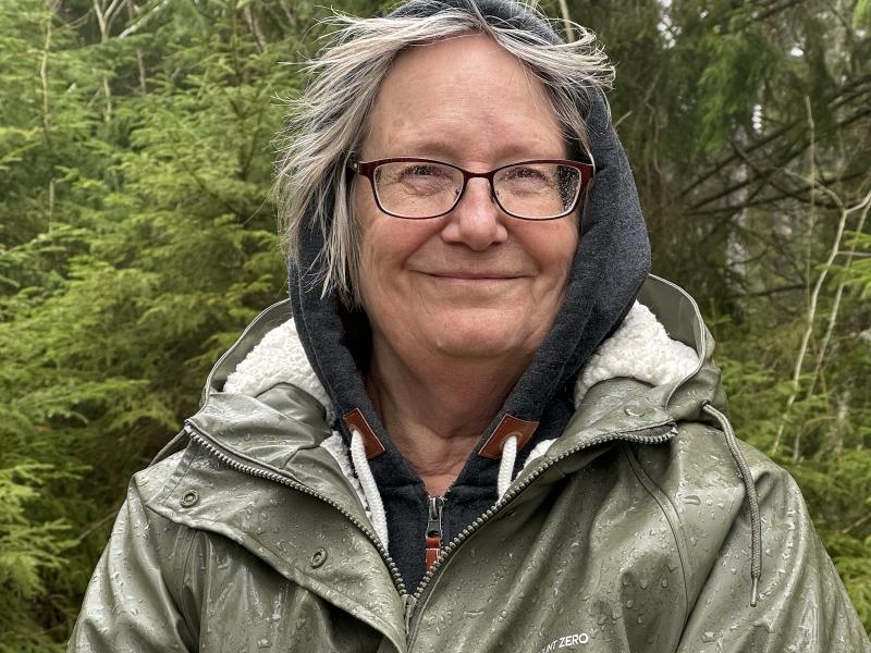 En äldre kvinna med grått hår och glasögon ler, klädd i en regnrock i en skogsmiljö.