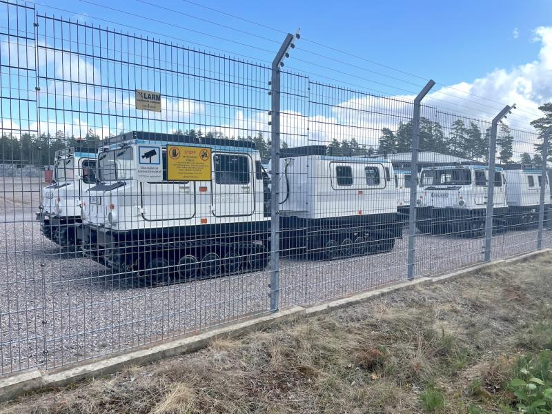 En rad vita militärfordon parkerade bakom ett högt vajerstängsel med skyltar som indikerar övervakning är närvarande.