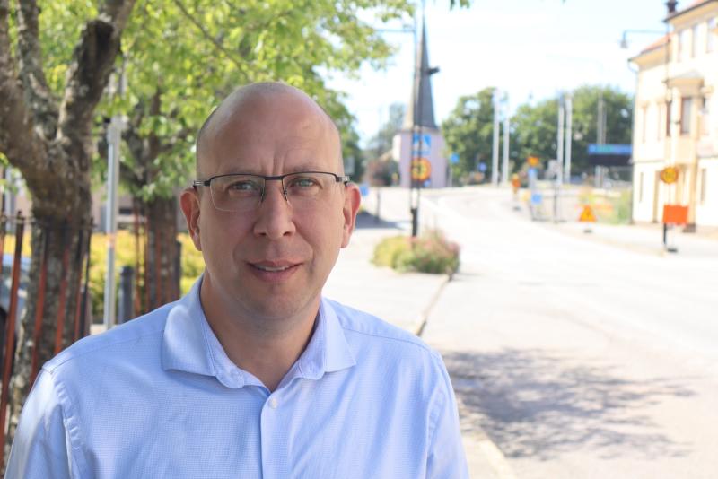 En man med glasögon och en vit skjorta står utomhus nära en lugn gata kantad av träd och byggnader en solig dag.