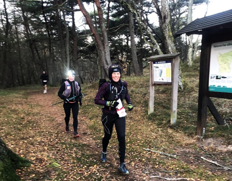 Löpare i en skog iklädda atletiska kläder, en med pannlampa, passerar en träskylt. Marken är täckt av löv.