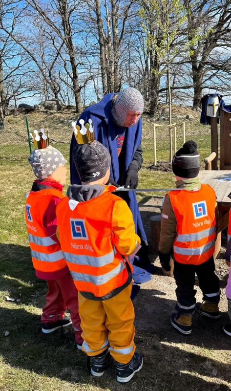 En vuxen i mössa pratar med tre små barn som bär reflexvästar och papperskronor utomhus.