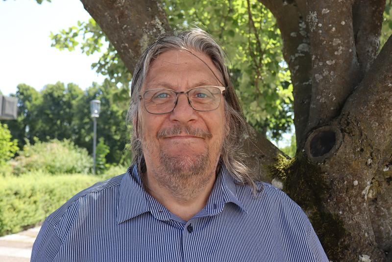 En äldre man med långt grått hår och glasögon står utomhus framför ett träd en solig dag, klädd i en randig skjorta, och ler svagt mot kameran.