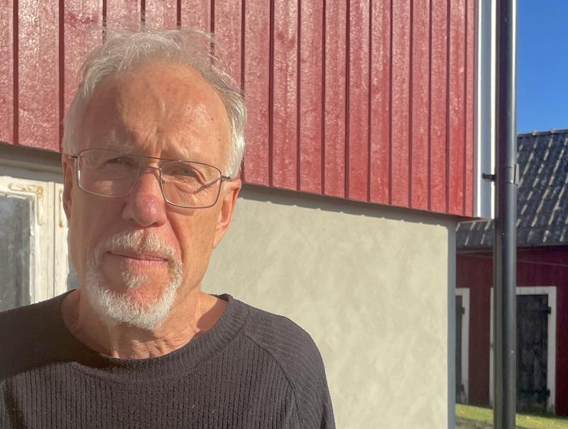 Äldre man med glasögon och skägg står i solljus framför en röd och grå byggnad.