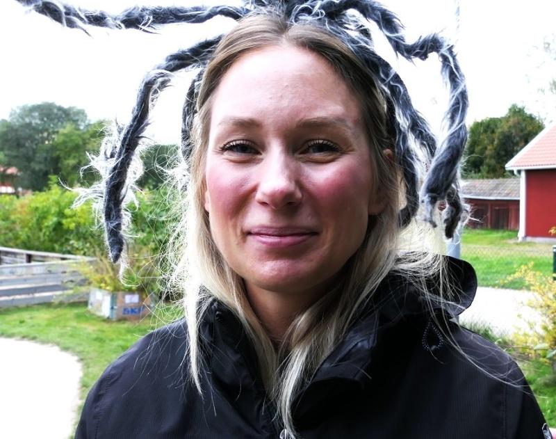 En kvinna i svart jacka ler utomhus samtidigt som hon bär ett pannband med stora, svartvita spindelben på.