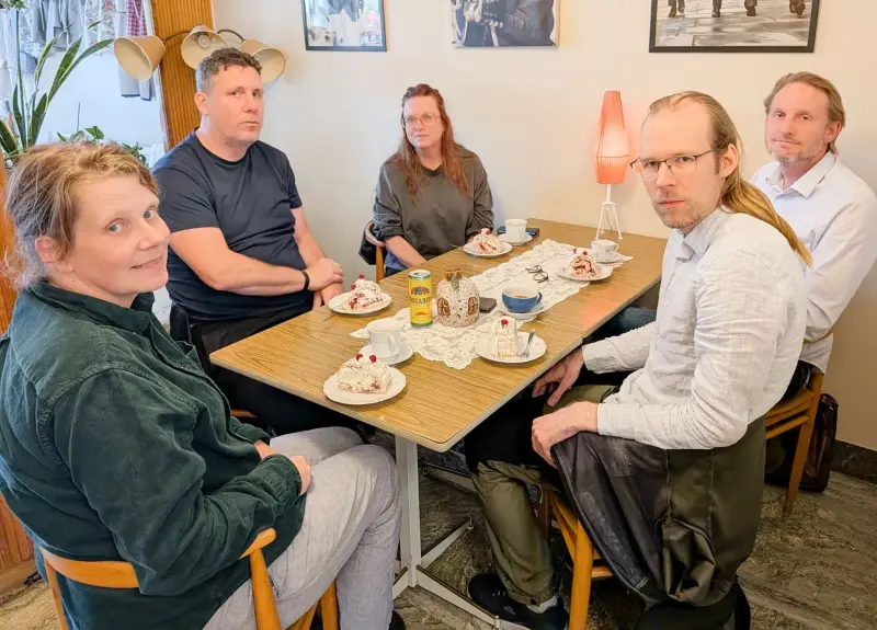 Fem vuxna sitter runt ett bord med desserttallrikar, koppar och en tekanna i ett mysigt rum med varm belysning och väggdekorationer.