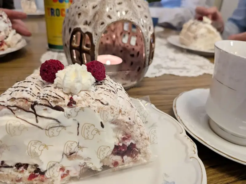 En bit gräddtårta med hallon och chokladglasyr ligger på en tallrik bredvid en kopp, med en ljusstake och människor i bakgrunden.