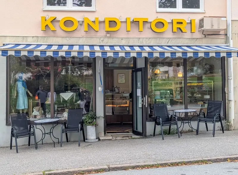 Ett bageri med en randig blåvit markis, en skylt med texten ”KONDITORI”, bord och stolar utanför samt tårtor utställda inne.