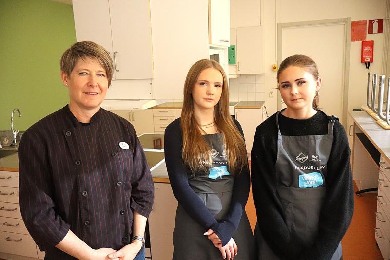 Tre kvinnor står i ett kök, två med förkläden och en i mörkrandig kockjacka, och tittar in i kameran.