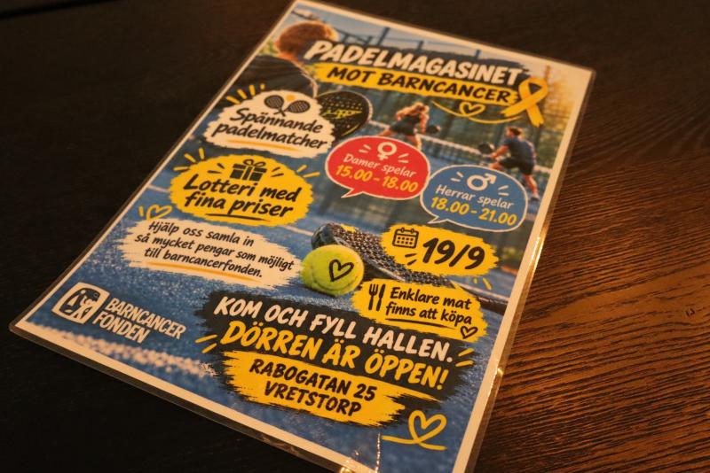 På ett bord ligger en flygblad som marknadsför en paddel-tävling till förmån för kampen mot barncancer, med matchtider, information om lotteriet och uppgifter om platsen på svenska.