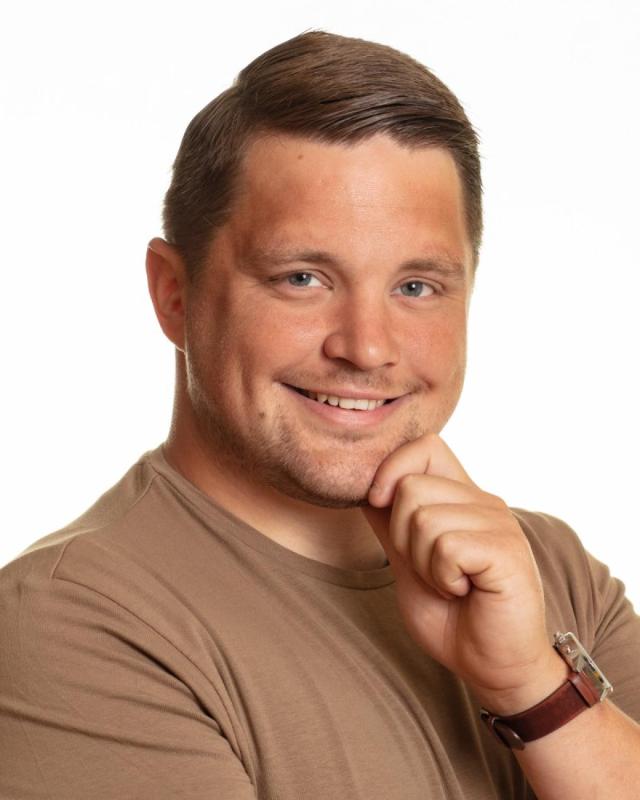 En person med kort hår iklädd beige skjorta och armbandsur ler och vilar hakan på handen.