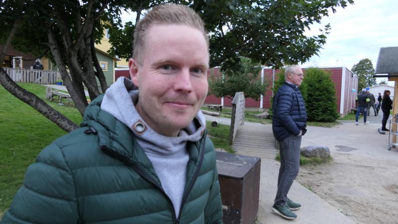 En man i grön jacka ler mot kameran i en utomhuspark; en annan person står i bakgrunden nära träd och byggnader.
