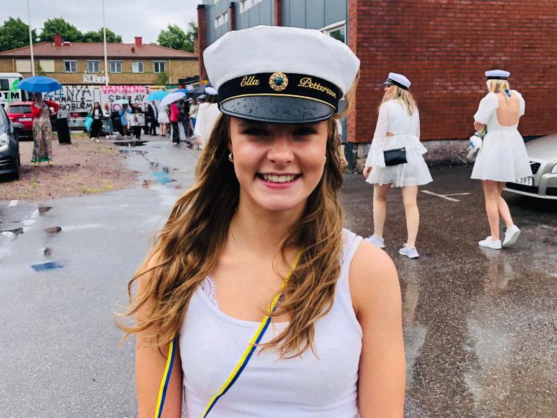 En flicka i vit outfit och examensmössa står utanför en regnig dag och ler mot kameran. Andra personer i liknande klädsel finns i bakgrunden.
