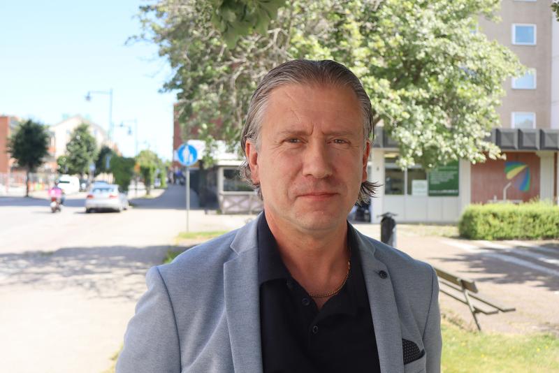 En man i en ljusgrå kavaj och svart skjorta står utomhus en solig dag, med träd och byggnader i bakgrunden.