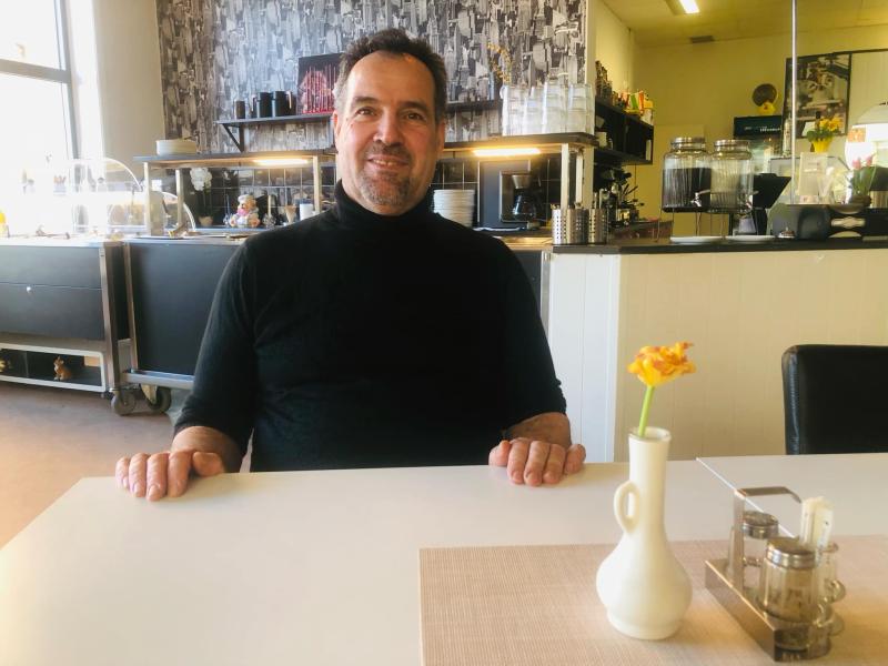 En man sitter vid ett bord på ett kafé, iklädd en svart skjorta. En liten vas med en gul blomma står på bordet och köksutrustningen syns i bakgrunden.