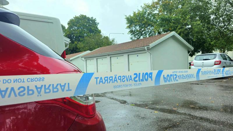 Polisens avspärrningsband blockerar tillträdet till ett parkeringsområde med garage och parkerade bilar en våt och molnig dag.