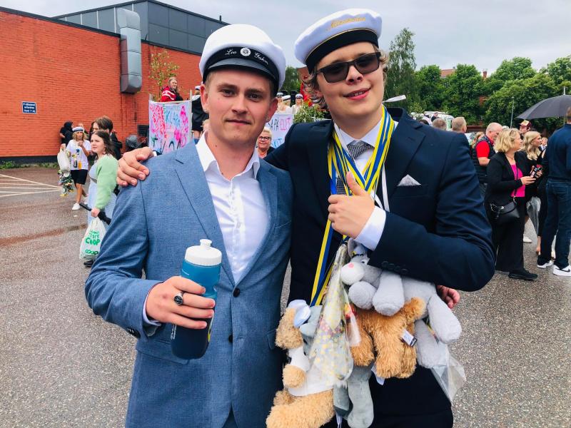 Två unga män i examensmössor och kostymer står bredvid varandra på en parkeringsplats. Den ene håller i en vattenflaska medan den andre bär många medaljer och gosedjur runt halsen.
