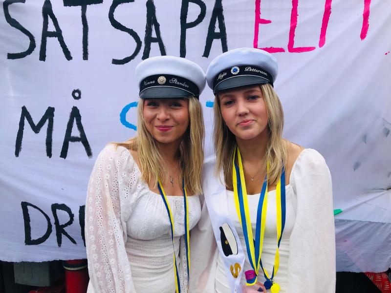 Två unga kvinnor i vita klänningar och studentkepsar står framför en banderoll med svensk text. Båda är prydda med gula och blå band.