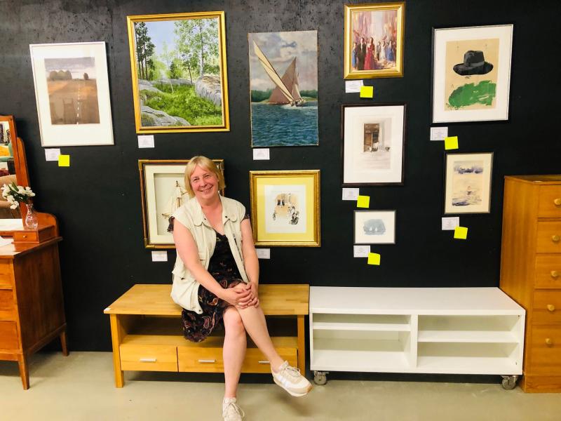 En kvinna sitter på en träbänk i ett galleri, omgiven av inramade konstverk på en svart vägg. Möbelbitar syns runt henne.