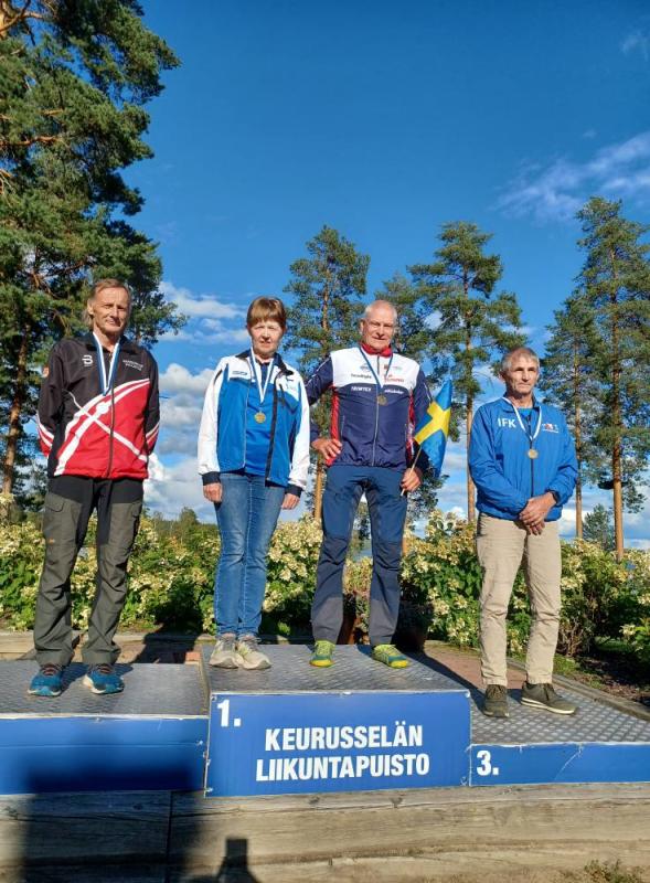 Fyra idrottare står på ett prispall utomhus under en blå himmel, iklädda medaljer. På podiet står det 