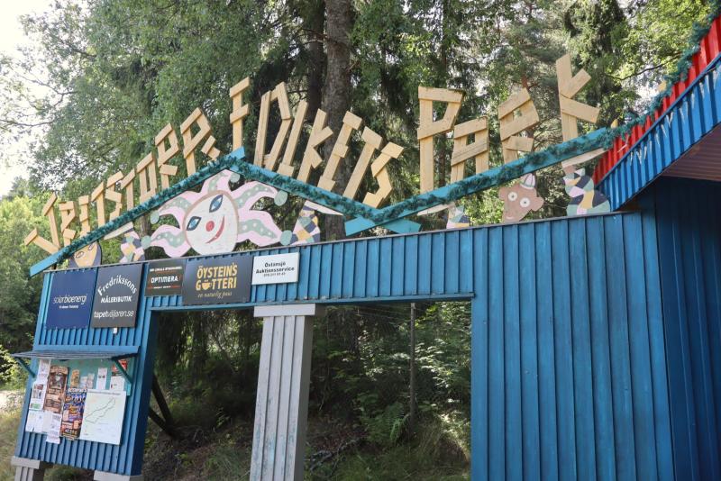 Ingången till ”Skrimser Fantasi Park” med färgglada, lekfulla skyltar och dekorationer, omgiven av gröna träd och ett blått trästaket.