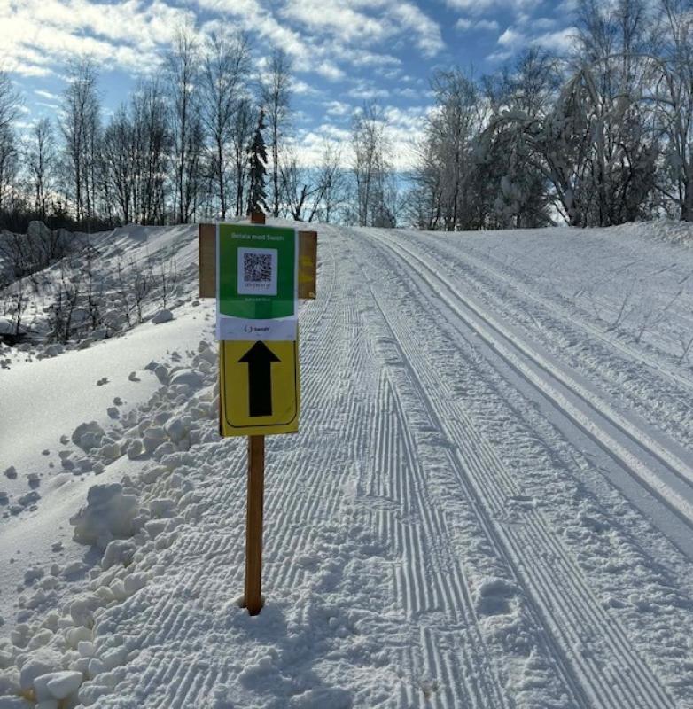 En spårskylt med en QR-kod och en stor svart pil pekar framåt på ett snötäckt längdskidspår under en delvis molnig himmel.