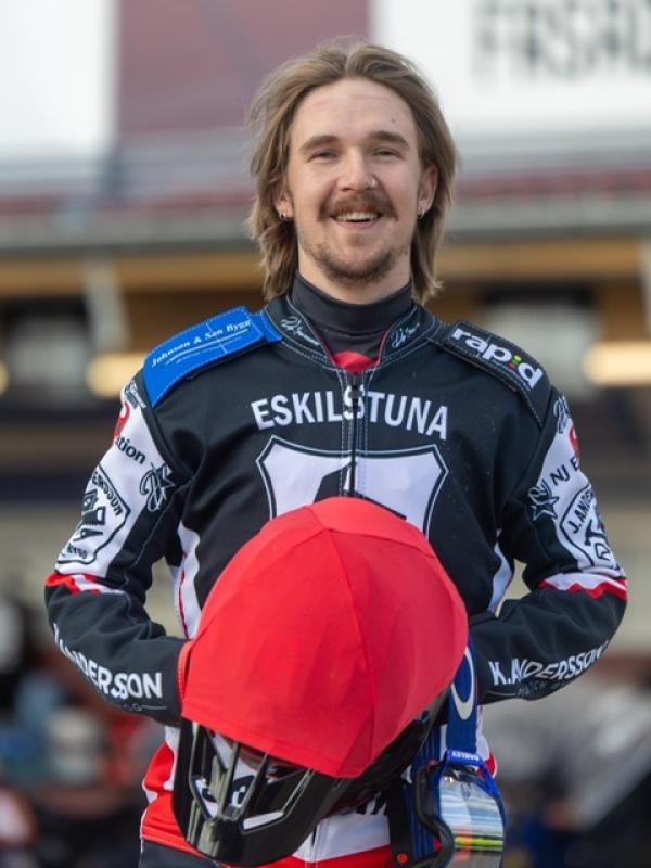 En speedwayförare i Eskilstunas lagdräkt håller i en röd hjälm och ler mot kameran, stående utomhus med suddiga byggnader i bakgrunden.