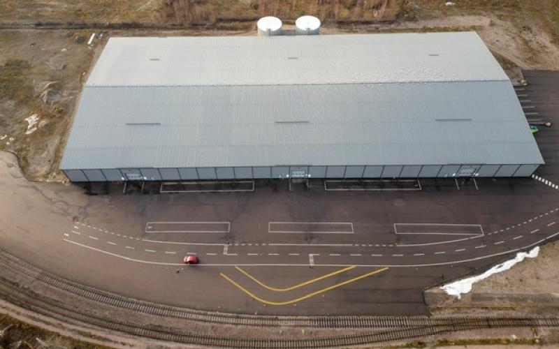 Flygfoto över en stor grå industribyggnad med en asfalterad tomt, tågspår i förgrunden och en röd bil parkerad utanför.