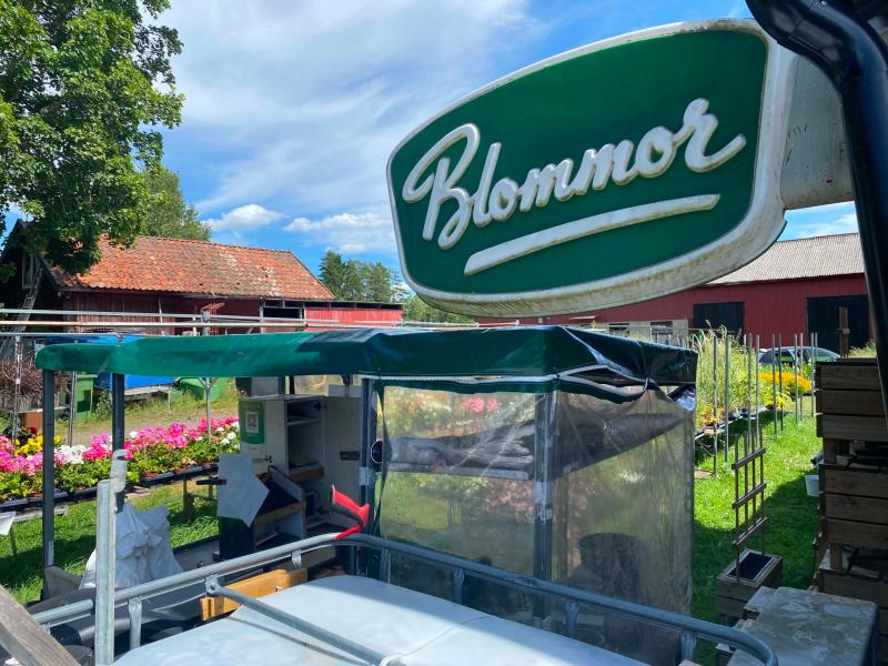 Ett blomsterstånd utomhus med en grön och vit skylt med texten ”Blommor”, krukväxter och trädgårdsinredning i en lantlig miljö med lador i bakgrunden.