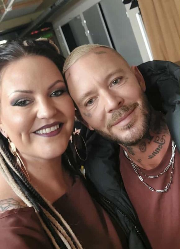 En kvinna med flätat hår och en man med ansiktstatueringar och ett kedjehalsband ler och poserar för en selfie tillsammans inomhus.