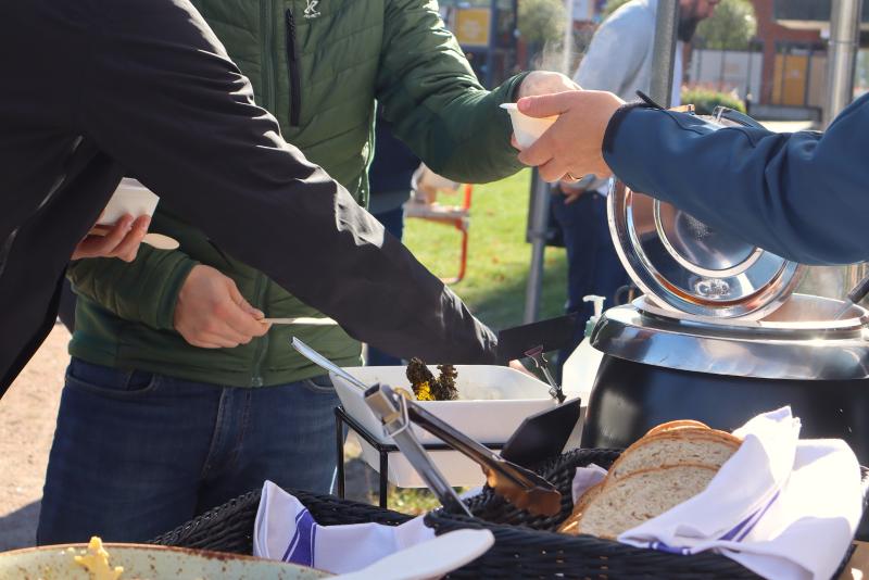 Tre personer serverar mat utomhus; en håller i en skål, en annan sträcker sig efter maten och en tredje står i närheten. På bordet syns bröd och soppa.