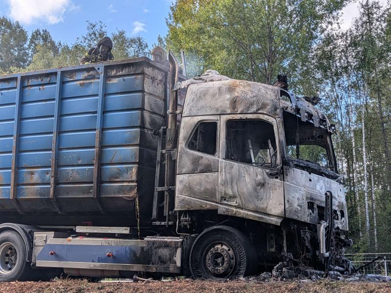 En stor blåvit lastbil med omfattande brandskador – med en förkolnad förarhytt och sotfläckar – står parkerad nära ett skogsområde.