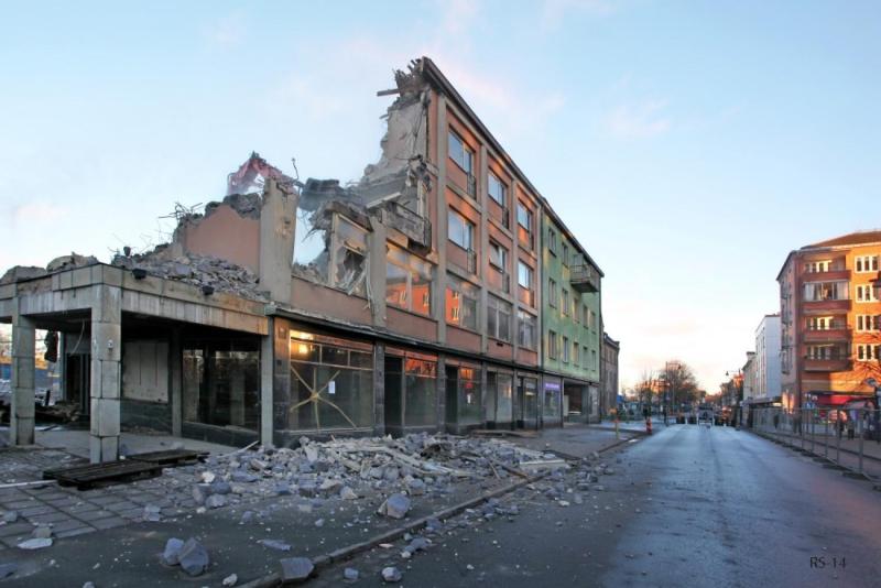 En delvis demolerad flervåningsbyggnad med bråte utspridd på trottoaren och en tom gata bredvid under en klar himmel.