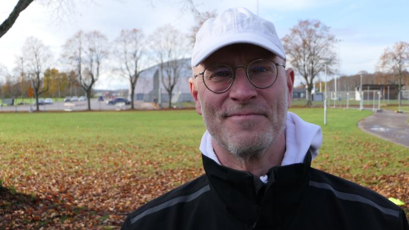 En man med glasögon, vit keps och svart jacka står utomhus på en gräsplätt med träd och nedfallna löv i bakgrunden.