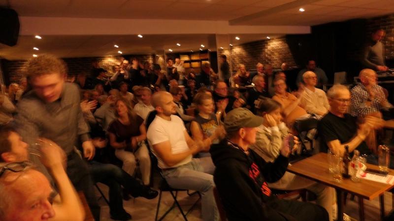 En stor grupp människor som sitter inomhus, applåderar och är vända framåt, med en livlig atmosfär i ett svagt upplyst rum.