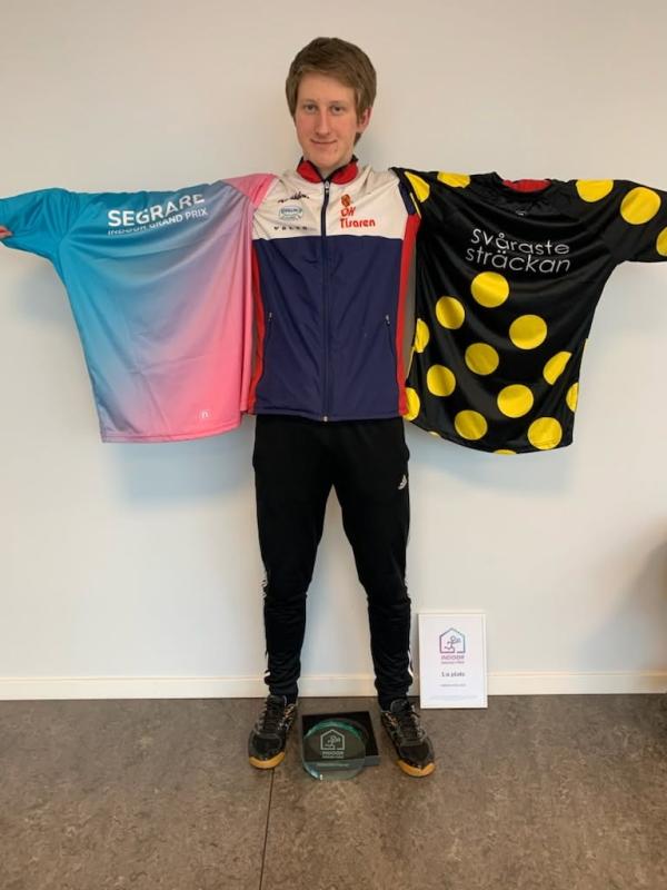 En person står mot en vägg och håller fram två cykeltröjor, en blå-rosa och en svart med gula prickar. En plakett och ett inramat certifikat ligger på golvet framför dem.