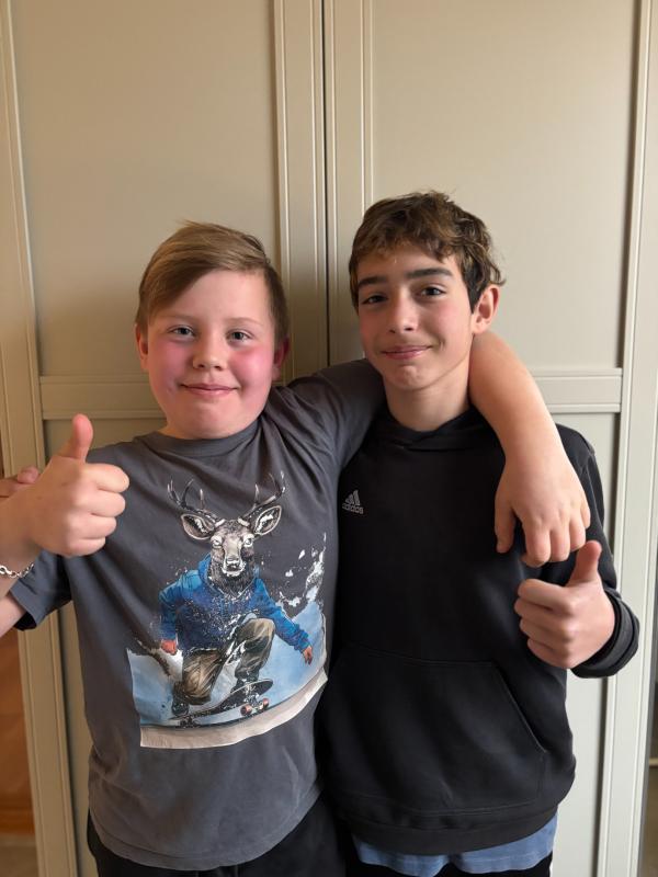 Två pojkar står sida vid sida inomhus och ler mot kameran. Pojken till vänster gör tummen upp och har på sig en T-shirt med en skateboardåkande hjort. Pojken till höger gör också tummen upp.