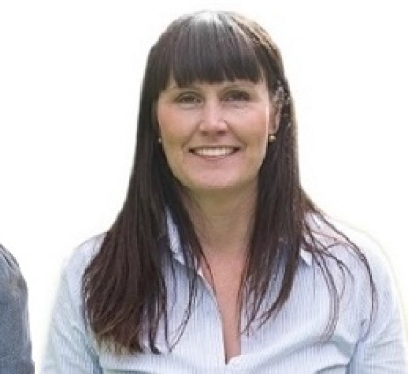 Leende person med långt mörkt hår klädd i en vit skjorta med knappar, stående mot en vanlig vit bakgrund.