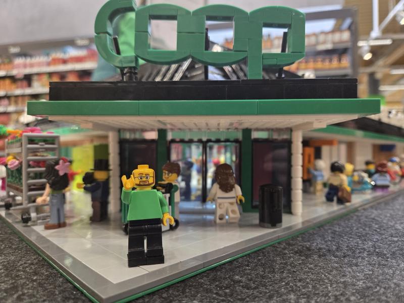 Legofigurer leker utanför en miniatyrbyggnad i grönt och svart som liknar en butik, där man kan se detaljerade hyllor och utställningar både inuti och i bakgrunden.