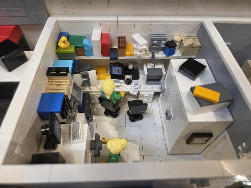 LEGO-figurer sitter vid skrivbord med datorer och kontorsmaterial i ett litet, detaljerat kontor byggt av LEGO-klossar, sett uppifrån.