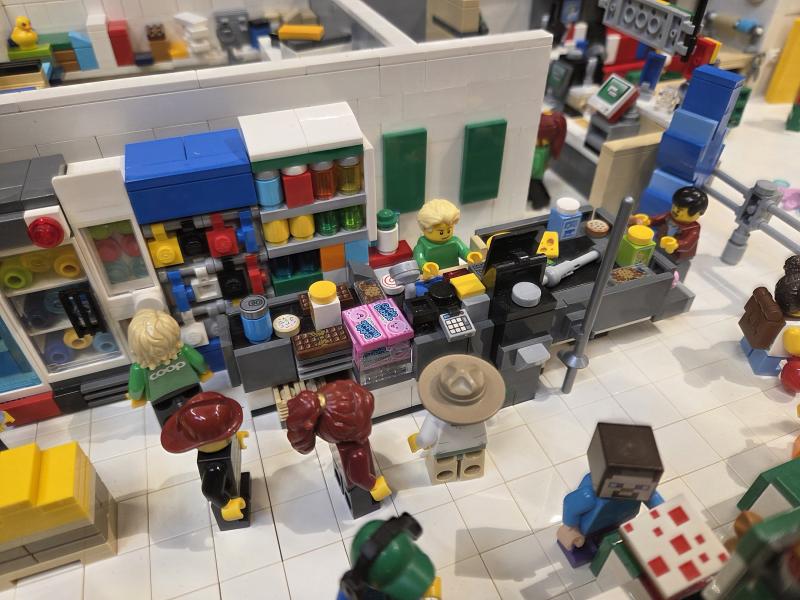Legofigurer står i kö vid en kassa i en stormarknad, medan en kassör skannar varor och hyllorna i bakgrunden är fyllda med olika varor.