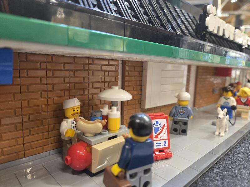 LEGO-figurer umgås vid en korvkiosk på trottoaren; en serverar mat, en annan står och väntar med en portfölj, och andra går förbi, däribland en figur med en hund.