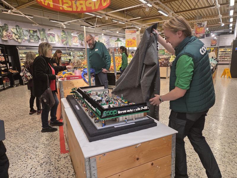 En person visar upp en detaljerad LEGO-modell av en stormarknad på ett bord inne i en livsmedelsbutik, medan flera personer betraktar utställningen.