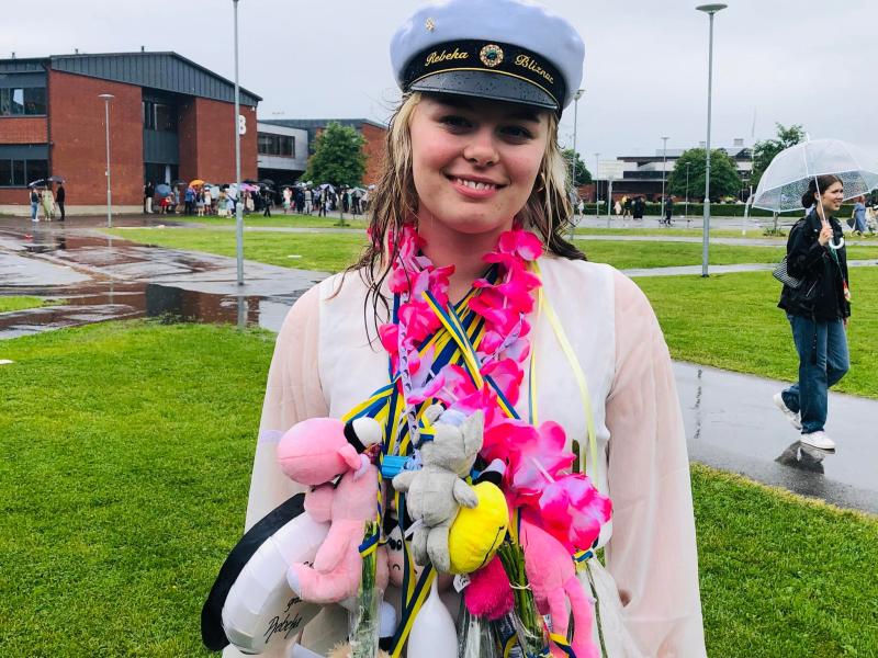 En ung person, iklädd vit keps och genomskinlig regnjacka, står på en gräsyta med en byggnad och människor i bakgrunden. De har flera stoppade leksaker och band runt halsen.