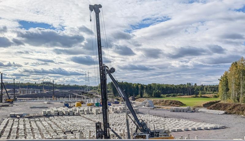 En byggarbetsplats med kranar, staplar av betongblock och maskiner under en delvis molnig himmel, omgiven av träd och gräsbevuxna fält.