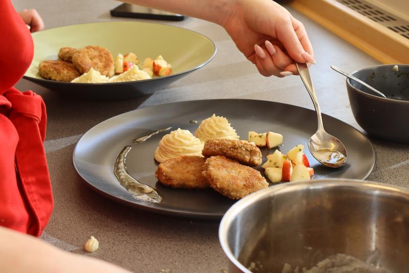 En person använder en sked för att lägga upp panerade kotletter, potatismos och små bitar av äpple på en svart tallrik. En annan tallrik med liknande mat ligger i närheten.