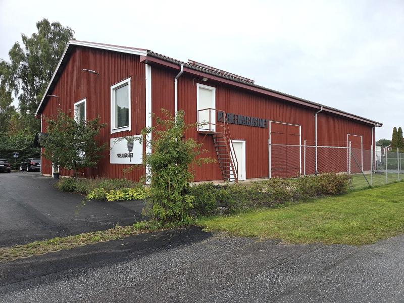 En röd träbyggnad med vita lister, en skylt med texten ”MEJERI MAGASIN” och en metalltrappa på utsidan, omgiven av grönska och ett stängsel av trådnät.