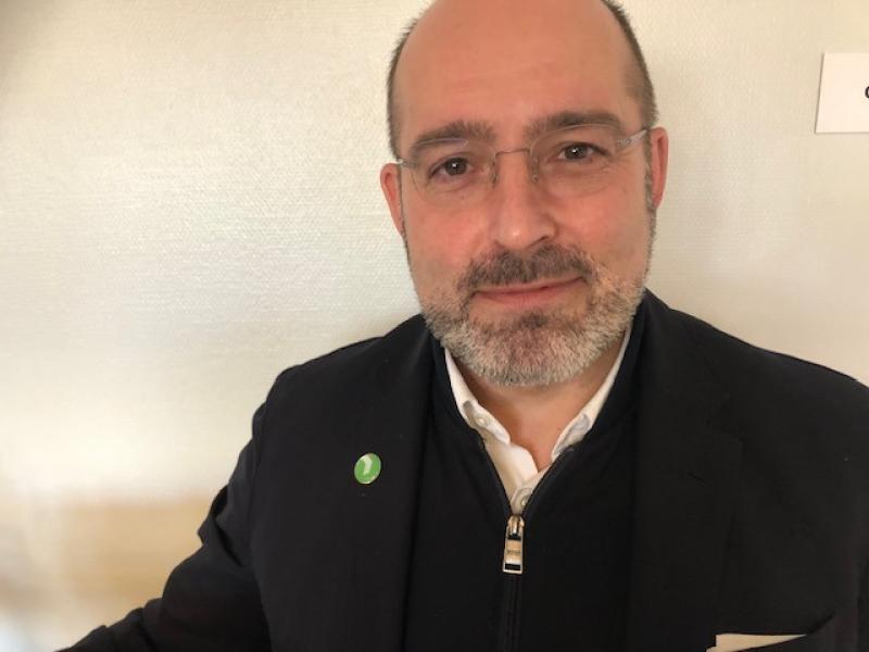 En medelålders man med glasögon och skägg, klädd i en mörk kavaj med en grön nål, står framför en ljus vägg.