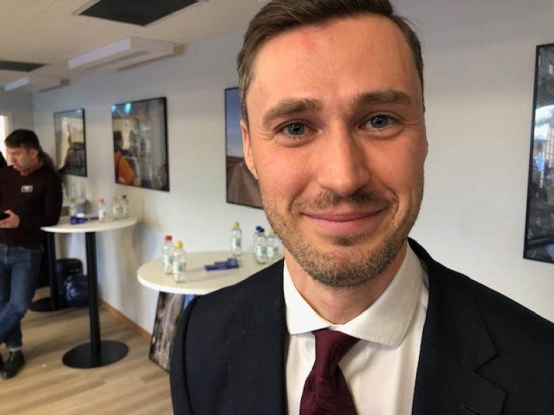 En man i kostym och slips ler mot kameran i ett modernt rum med små runda bord och vattenflaskor. Andra människor syns i bakgrunden.